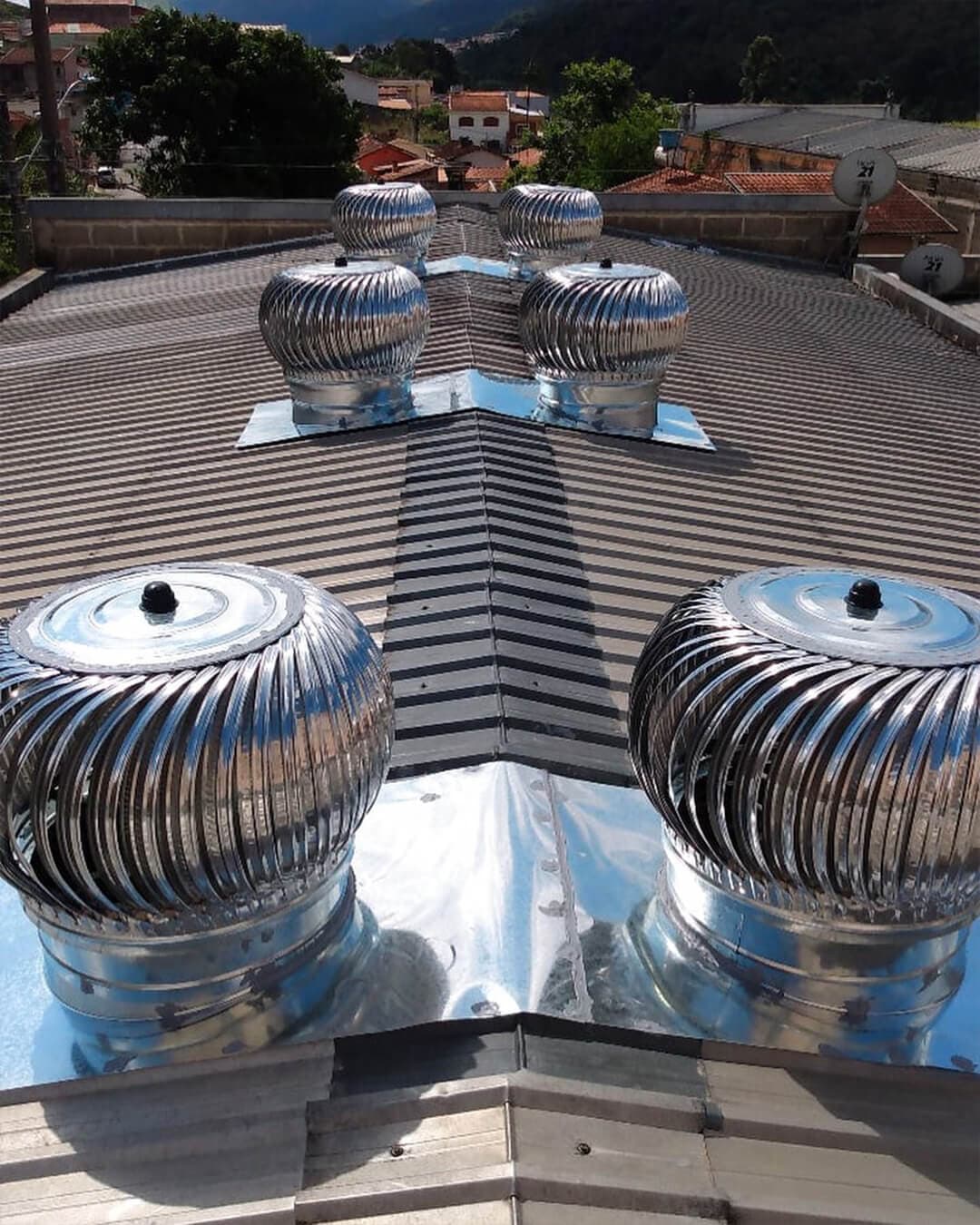 Exaustores eólicos de aço inox instalados em telhado metálico de galpão, com casas e montanhas ao fundo