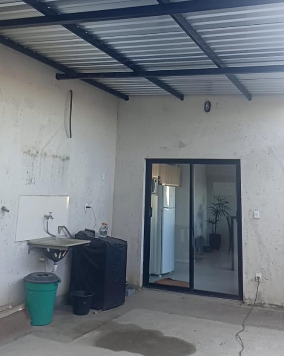 Cobertura metálica com telhas e vigas de aço sobre área de serviço externa, ao lado de porta de vidro com esquadria preta