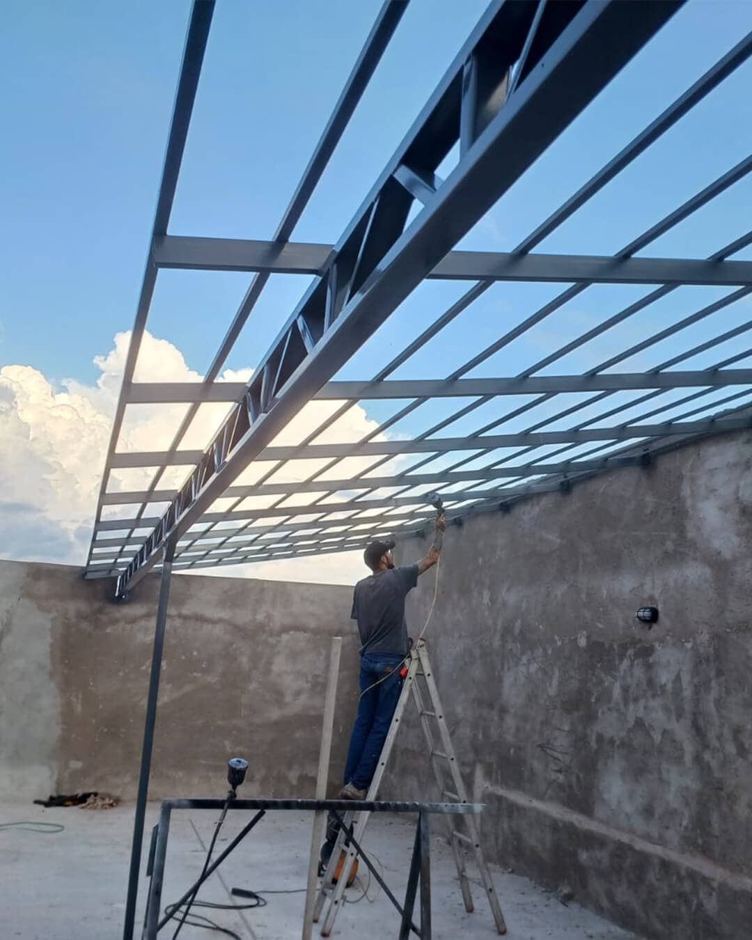 Profissional em escada aplicando pintura em estrutura metálica de cobertura sobre paredes de alvenaria em obra