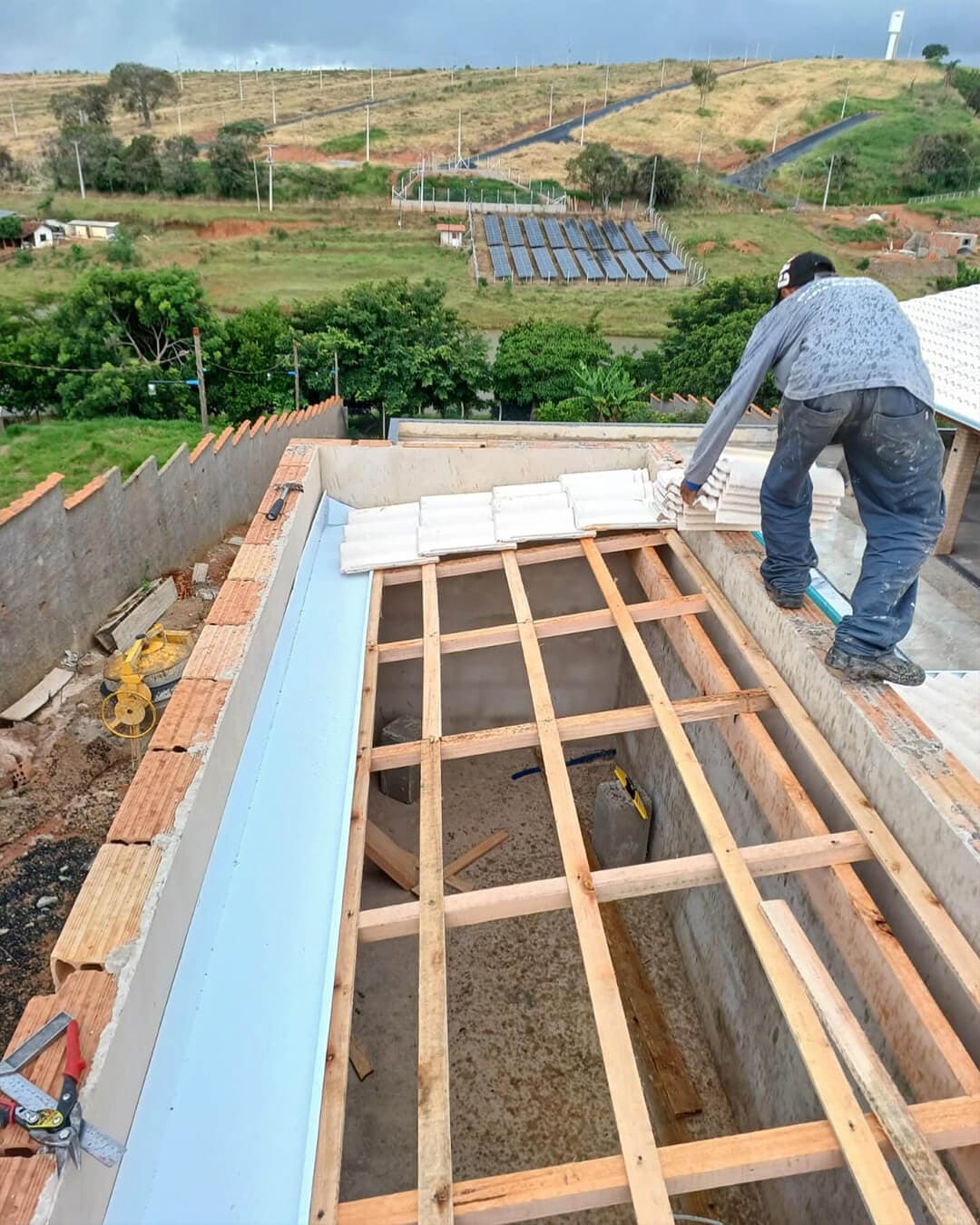 Instalação de calha metálica e madeiramento de telhado em obra residencial, com profissional posicionando telhas na laje
