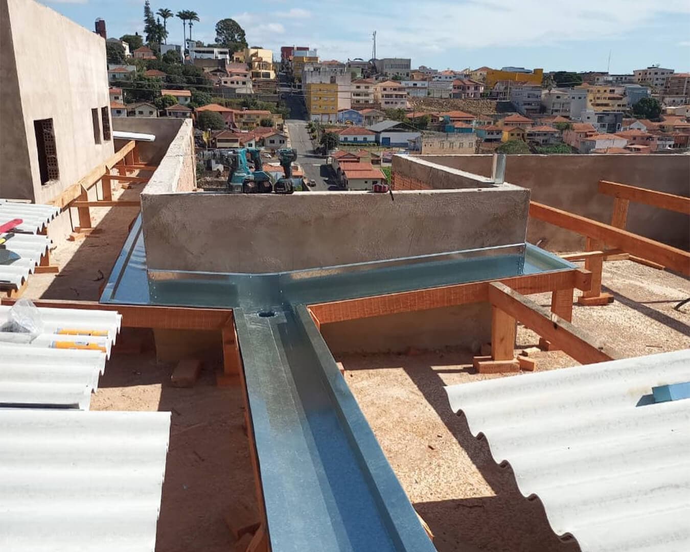 Calha metálica galvanizada instalada em telhado em construção, com telhas de fibrocimento e casas ao fundo